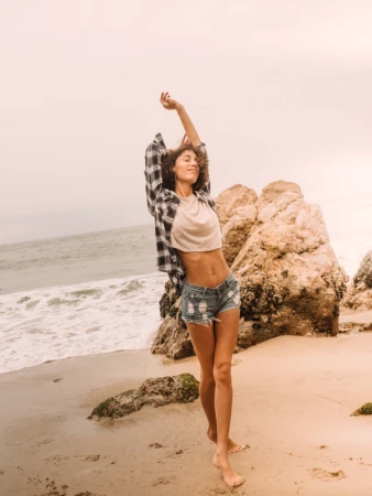 Woman stretches on a sandy beach with a flannel shirt, crop top, and distressed denim shorts, enjoying a serene ocean view on a cloudy day.