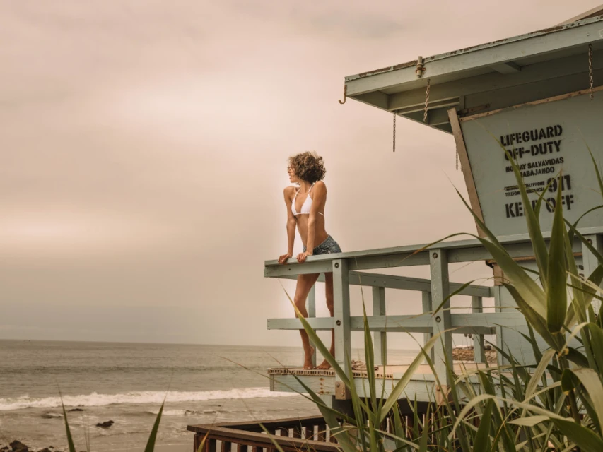 A woman in a bikini and denim shorts stands on a lifeguard tower overlooking the ocean on a cloudy day at the beach.