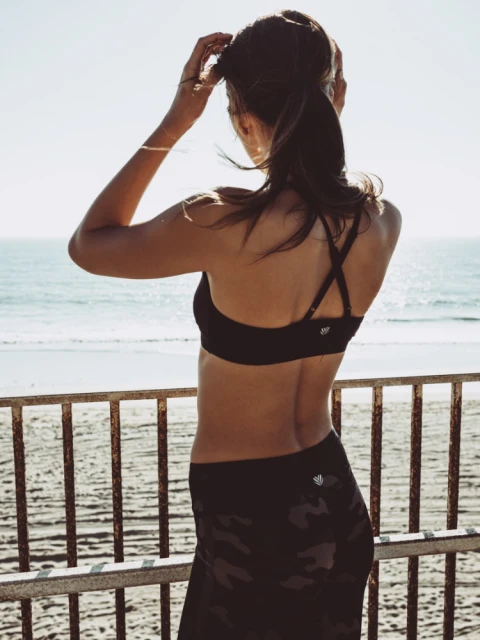 Woman in sports bra and camouflage leggings standing on beach. Athletic female facing ocean with blue sky backdrop and metal railing.