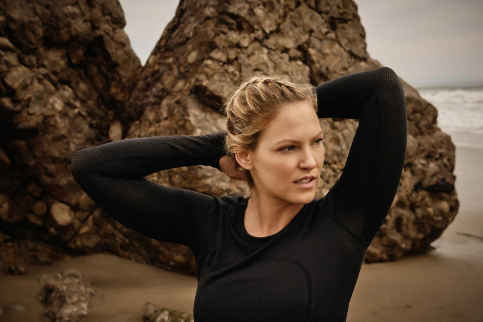 Blonde woman in athletic wear poses against a large rock on a beach, with waves visible in the background on an overcast day.