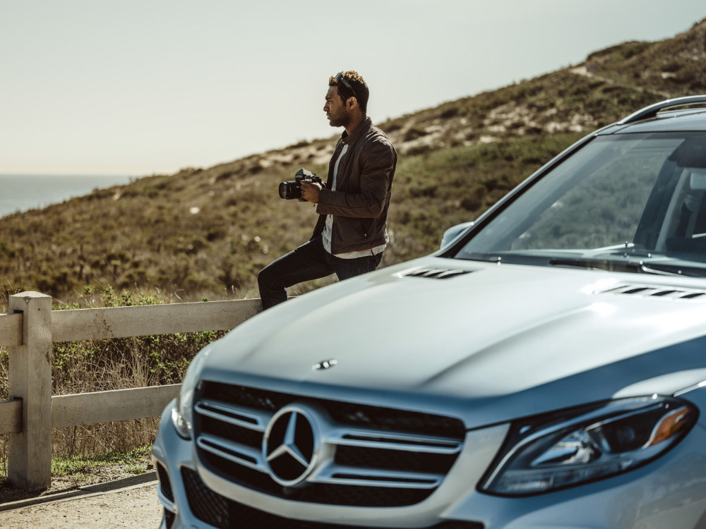 Man sits on railing with camera near silver Mercedes-Benz SUV overlooking ocean on sunny day; outdoor automotive lifestyle photoshoot.
