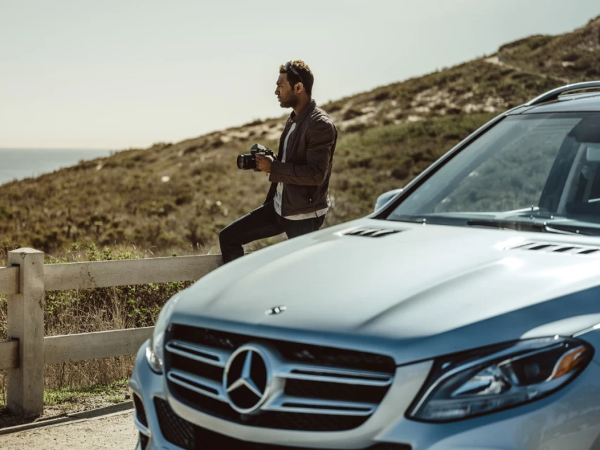 Man sits on railing with camera near silver Mercedes-Benz SUV overlooking ocean on sunny day; outdoor automotive lifestyle photoshoot.