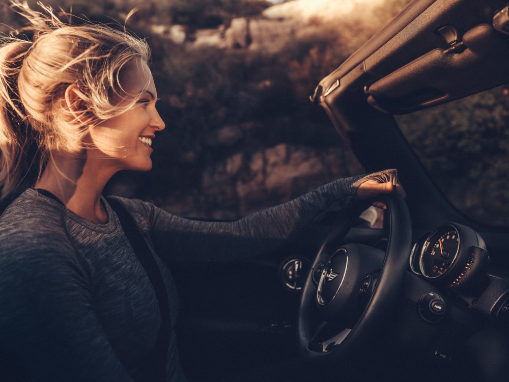 Blonde woman smiles while driving a convertible car in an outdoor setting with trees in the background, illuminated by sunlight.