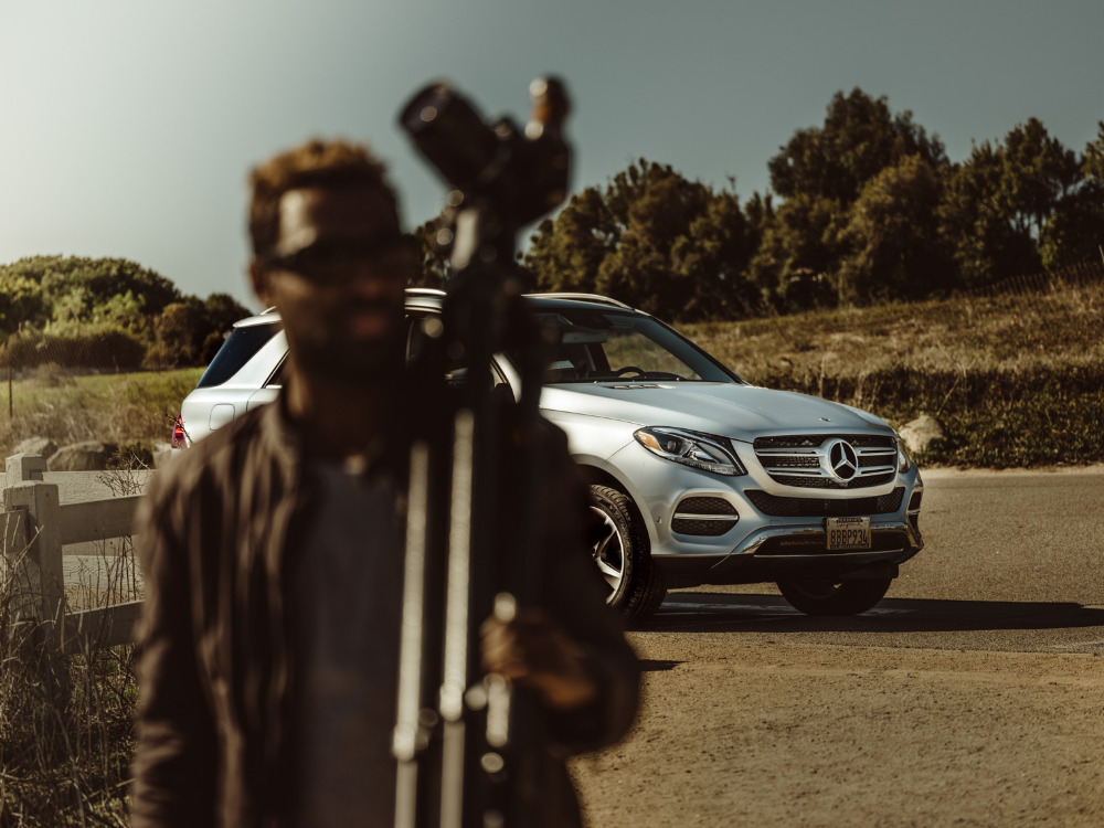 Silver Mercedes-Benz GLE SUV parked on an outdoor road with a blurry adult male holding a camera tripod in the foreground.