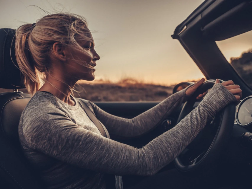 Blond woman driving a convertible car at sunset, wearing a grey top and seatbelt, wind blowing through her hair on a road trip.