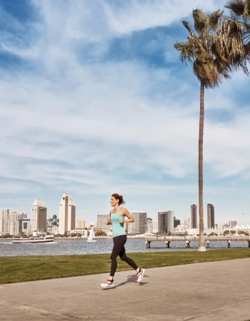 Woman jogging along a waterfront path with tall palm trees and a city skyline across the water in the background.