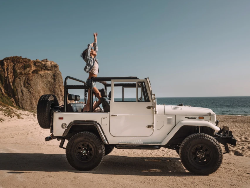 A woman stands atop a white Jeep Wrangler on a beach with a rocky cliff and ocean view under a clear blue sky, capturing a moment of freedom.