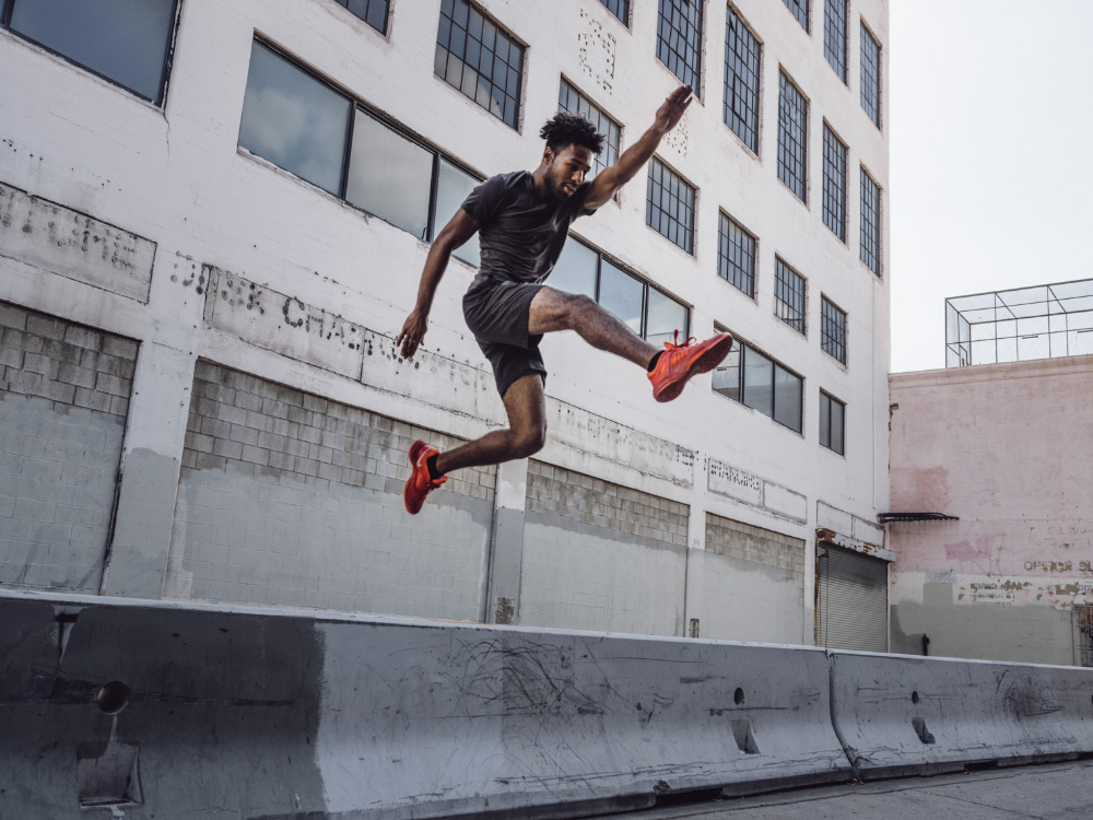 Athletic man jumps over concrete barrier in urban setting. He is wearing a dark tee shirt and shorts and bright orange athletic shoes.