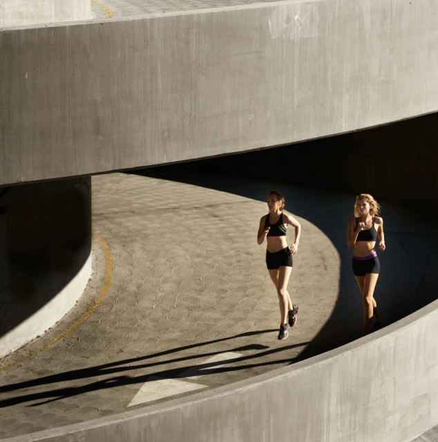Two adult women in athletic wear are running outdoors along a concrete ramp with dynamic shadows during daylight hours, fitness concept.
