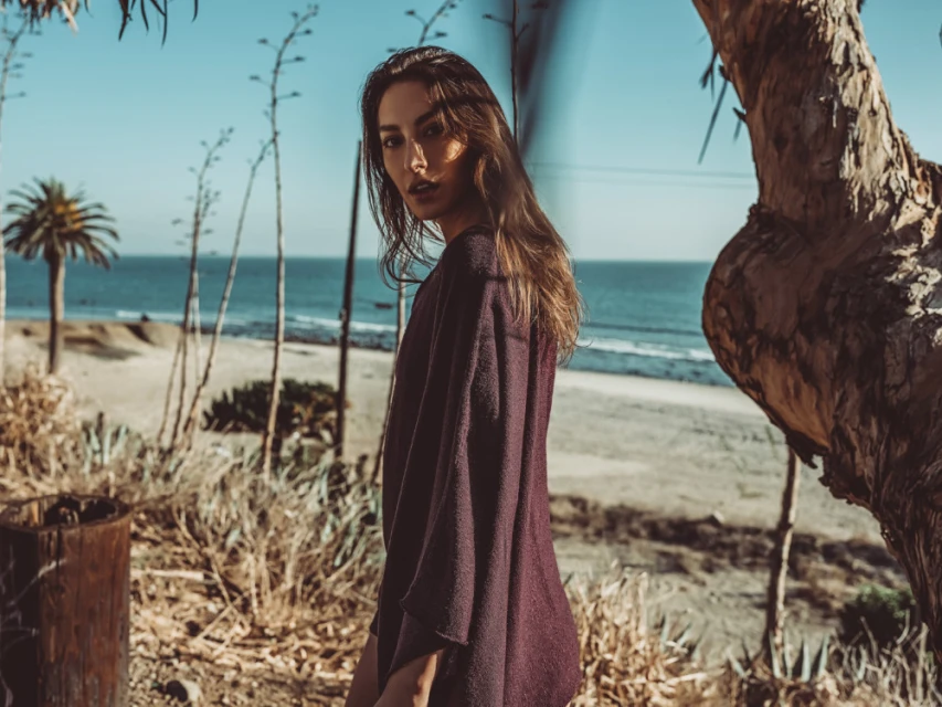 Adult female in a poncho stands near ocean, palm trees, and foliage on a sunny day, showcasing natural beauty and a relaxed outdoor lifestyle.