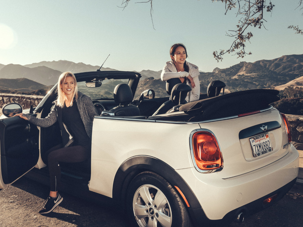Two women sit in a white convertible car with a mountain range in the background on a sunny day, with one woman exiting the car.