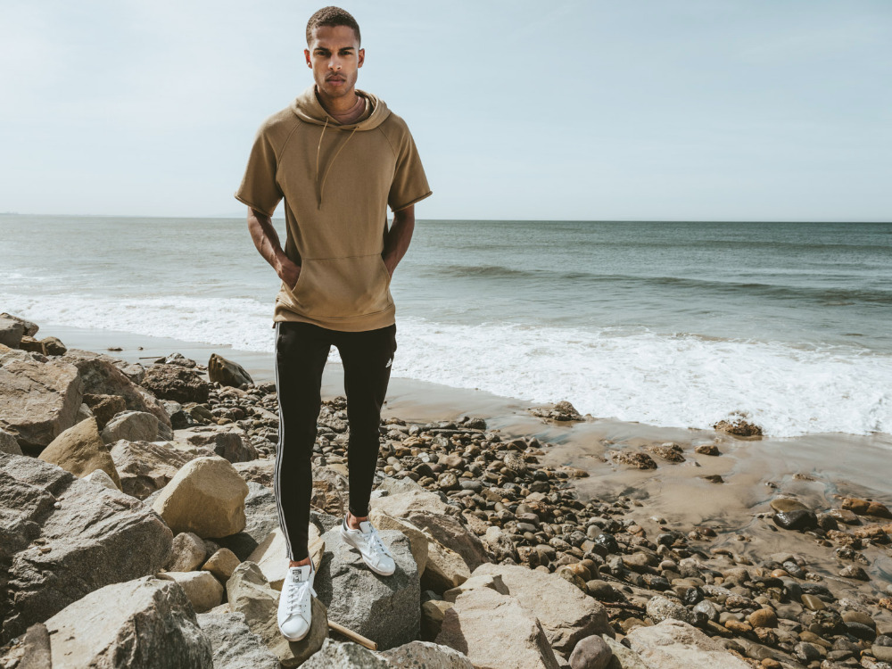 Young adult male stands on rocky beach near ocean in athletic wear, including a short-sleeve hoodie, track pants, and white sneakers.