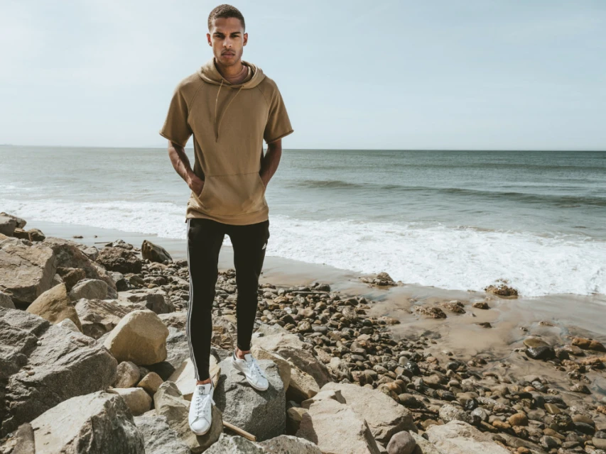 Young adult male stands on rocky beach near ocean in athletic wear, including a short-sleeve hoodie, track pants, and white sneakers.