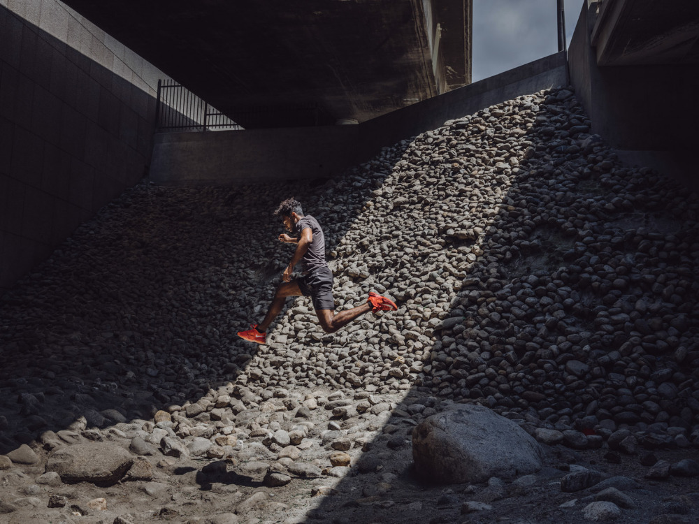 Adult male athlete in sportswear running uphill on a rocky slope under a bridge with dappled sunlight, showcasing fitness and determination.