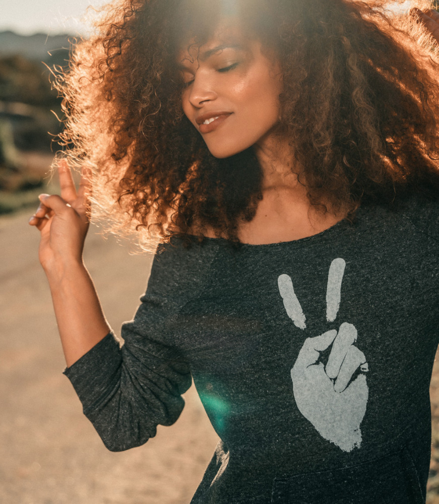 Woman with voluminous curly hair wearing a dark grey sweatshirt with a white peace sign graphic outdoors.
