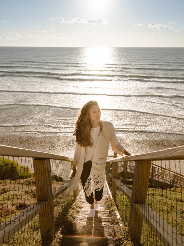 Woman descends wooden stairs towards sunlit ocean view. She wears a cream shawl, dark pants, white top, creating a serene coastal scene.
