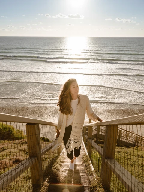 Woman descends wooden stairs towards sunlit ocean view. She wears a cream shawl, dark pants, white top, creating a serene coastal scene.