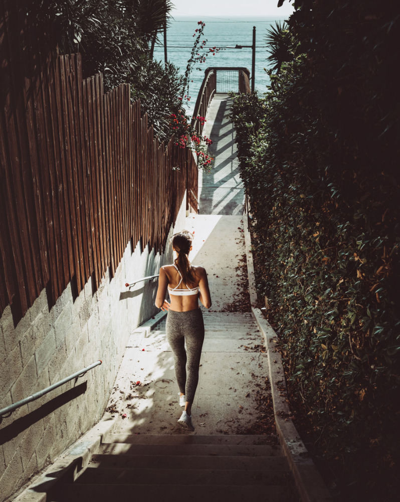 Woman in sports bra and leggings ascends outdoor stairs towards the ocean. Lush green foliage lines the stairwell on a sunny day.