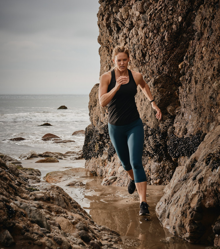 A woman running between rock formations on a beach with the ocean in the background, captured in a medium shot with natural lighting.