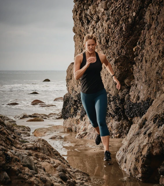 A woman running between rock formations on a beach with the ocean in the background, captured in a medium shot with natural lighting.