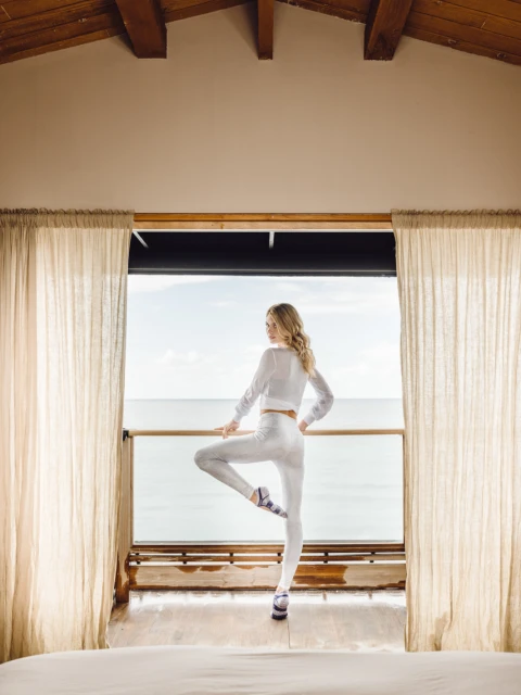 Young woman in white sportswear does a yoga tree pose on a balcony overlooking the ocean with cream drapes and wooden ceiling.