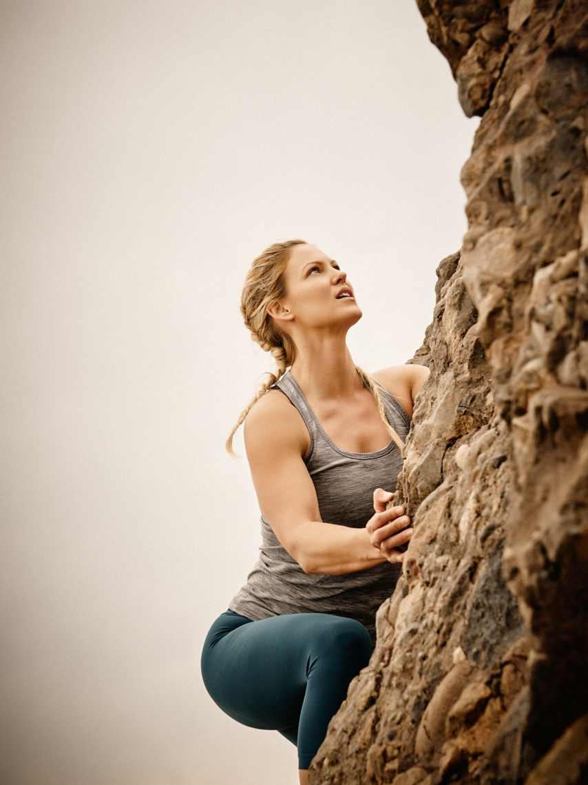 A woman in athletic wear is rock climbing with a determined expression against a neutral sky background in an eye-level shot.