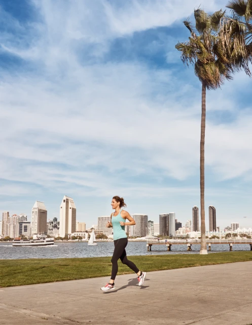 A woman in fitness attire runs along a waterfront path in San Diego, with the city skyline, palm trees, and ocean in the background.