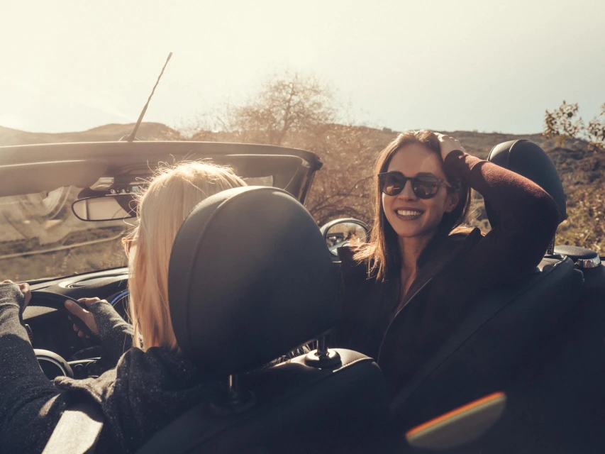 Two smiling women in sunglasses enjoy a sunny road trip in a convertible car through a natural landscape with hills and sparse vegetation.