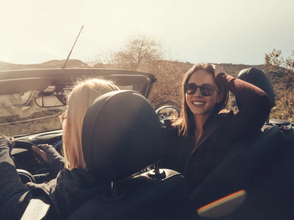 Two smiling women in sunglasses enjoy a sunny road trip in a convertible car through a natural landscape with hills and sparse vegetation.