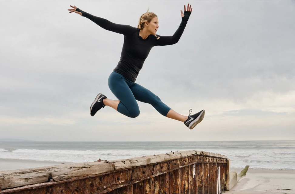 Athletic woman in sports wear leaps off a weathered pier with outstretched arms, ocean waves crashing behind against a cloudy sky.