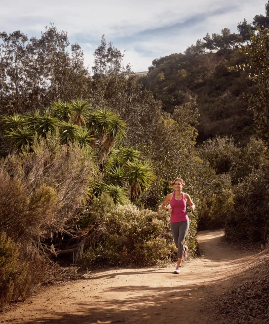 Woman in sportswear runs on a dirt path surrounded by lush green trees and bushes in a sunny outdoor fitness setting.