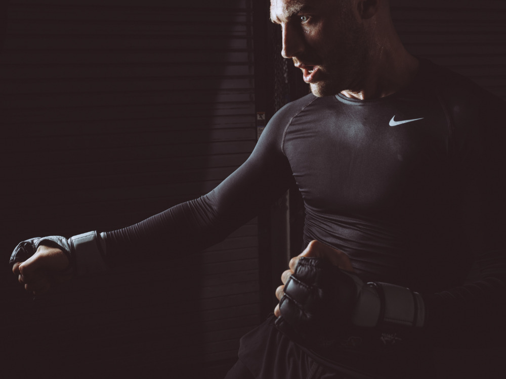 A muscular man is boxing in a dimly lit room, wearing black gloves and a long-sleeved shirt featuring a visible Nike logo, showcasing intense sport and fitness.