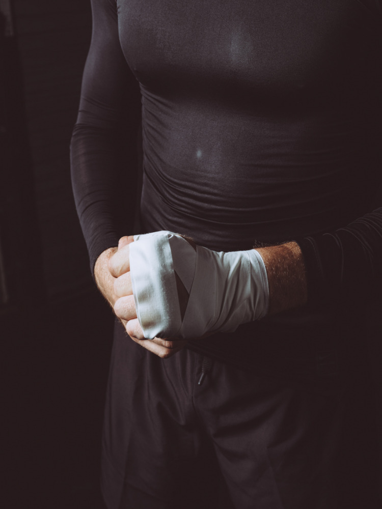 Close-up shot of a man wrapping his hands with white bandages, wearing a black long-sleeved shirt, against a dark background.