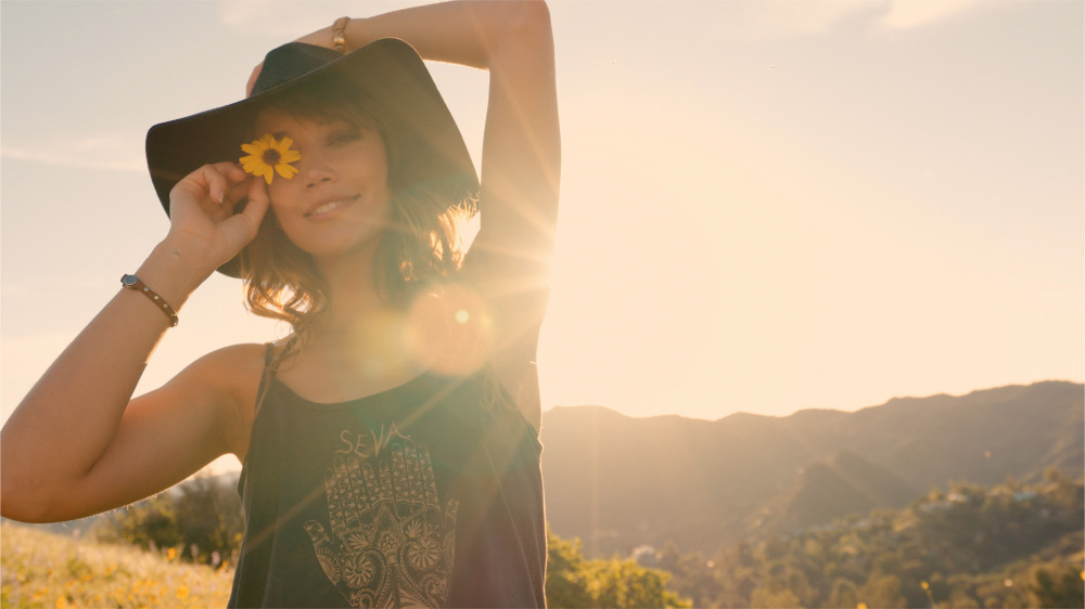 Young woman with a tan holds a yellow flower to her eye, wearing a black floppy hat in a sunny, outdoor field with mountains in background.