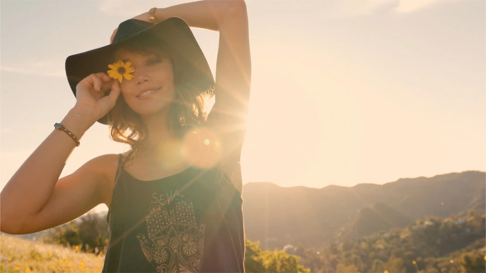 Young woman with a tan holds a yellow flower to her eye, wearing a black floppy hat in a sunny, outdoor field with mountains in background.