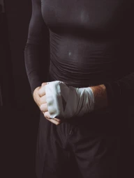 Man wearing a black athletic top wrapping his hands in white tape against a dark background