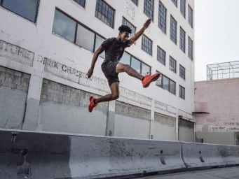 Black man mid-air jumping over a concrete barrier wearing dark workout clothes and bright red sneakers