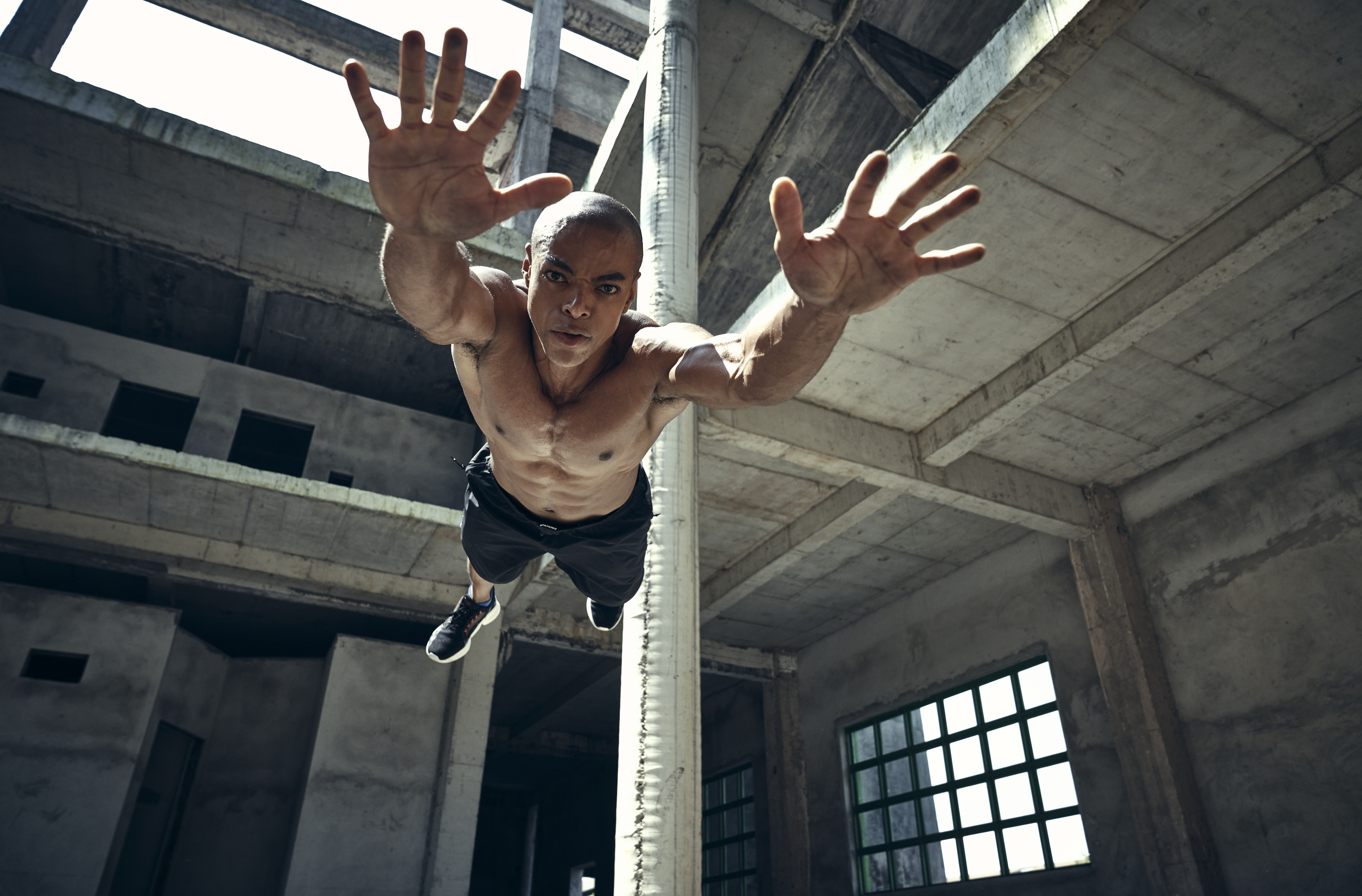 Muscular shirtless man mid-air in an abandoned building reaching forward, low-angle shot emphasizing urban decay and strength.