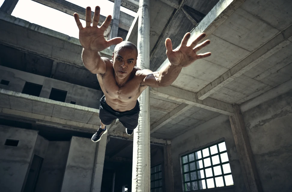 Muscular shirtless man mid-air in an abandoned building reaching forward, low-angle shot emphasizing urban decay and strength.