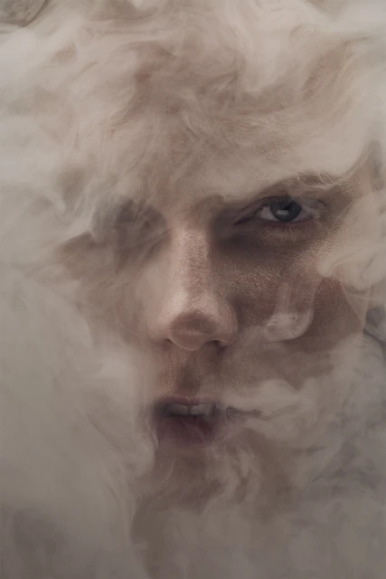 Dramatic close-up of a person's face partially obscured by swirling smoke, emphasizing expressive eyes and intense atmosphere in a studio.