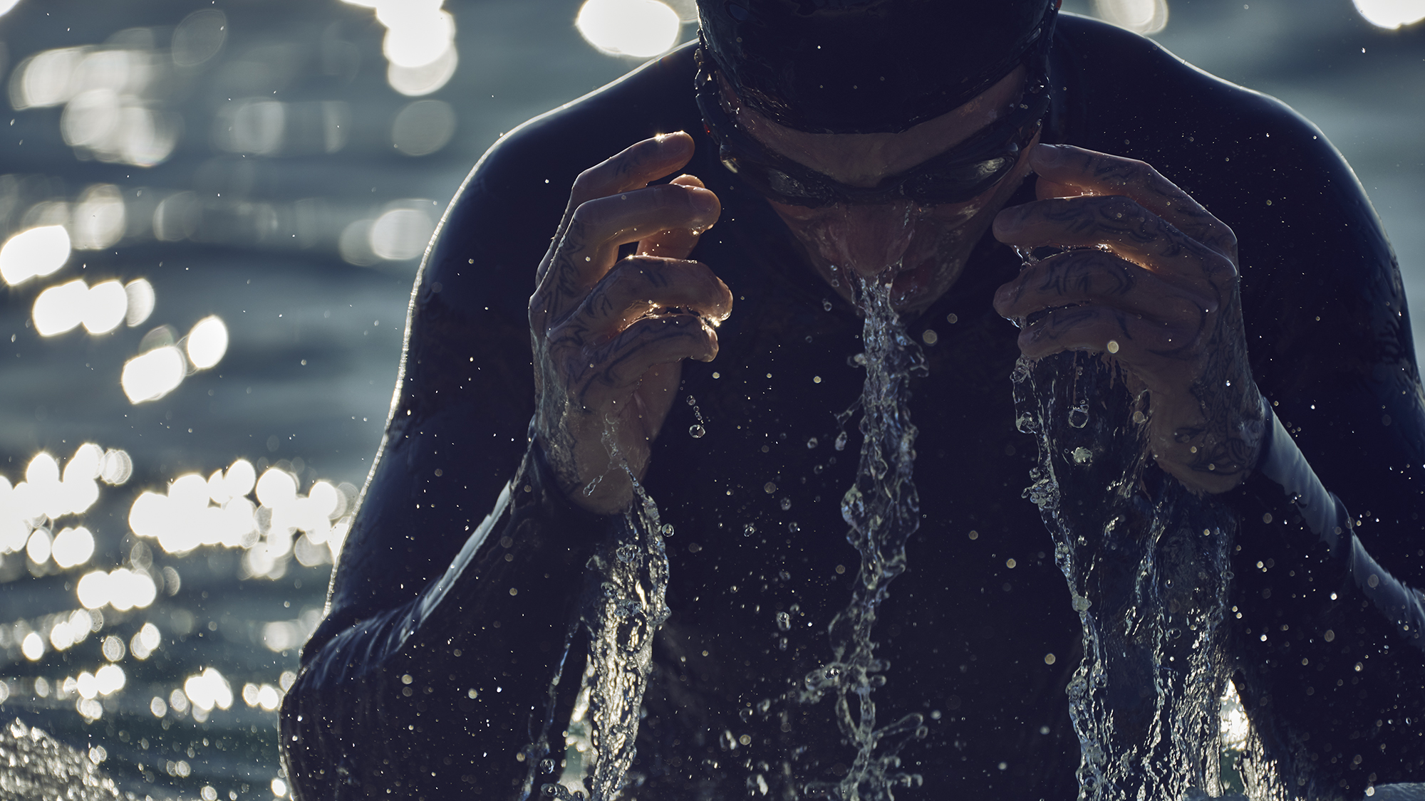 Adult male swimmer in wetsuit and goggles emerges from water, washing face, with bokeh water background and backlight.
