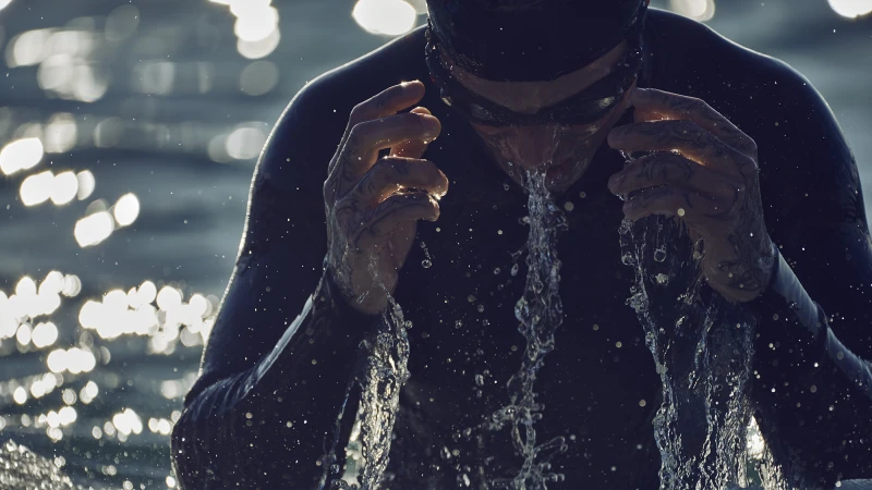 Adult male swimmer in wetsuit and goggles emerges from water, washing face, with bokeh water background and backlight.