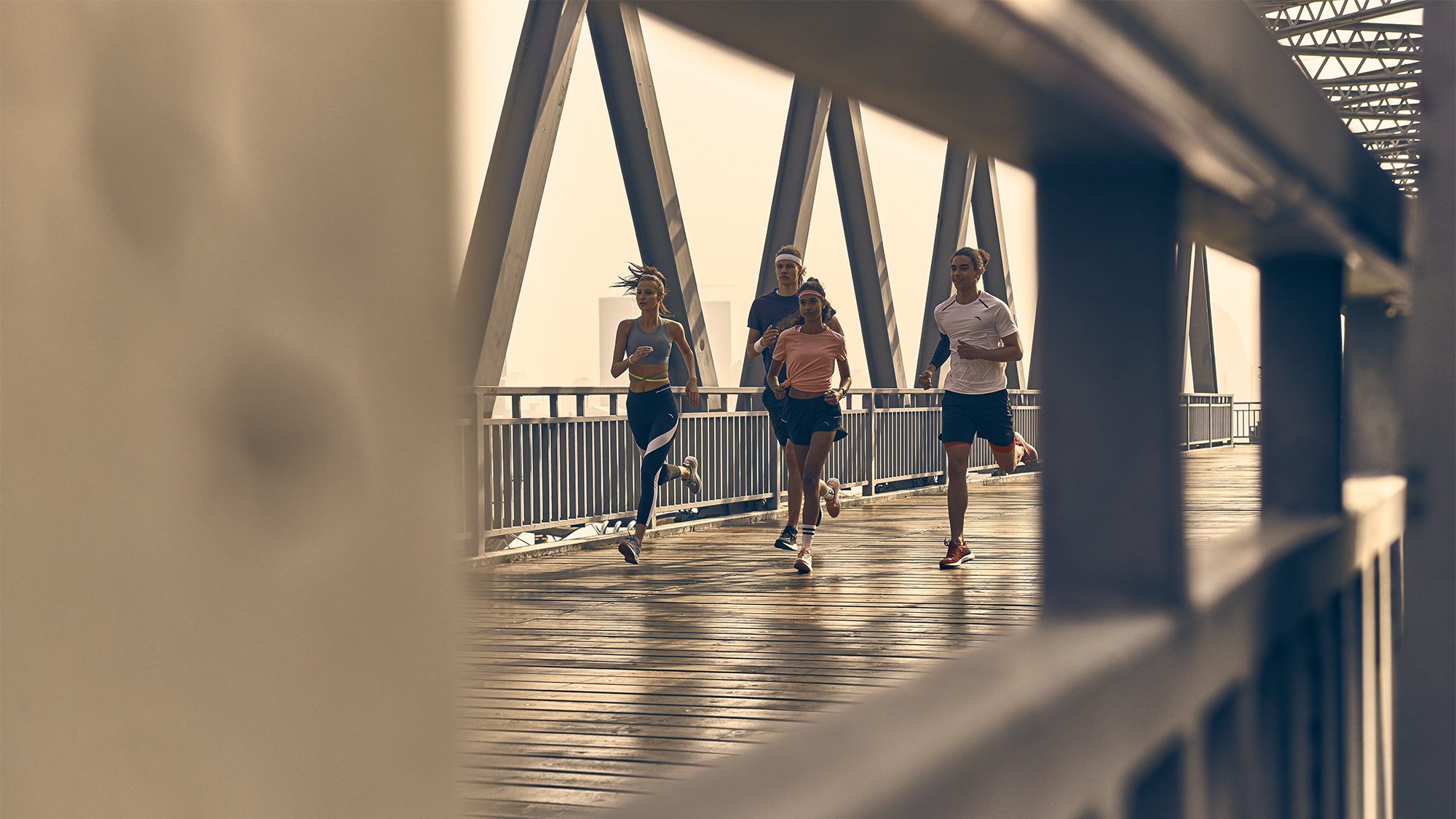 Four people are running on a metal bridge in sportswear. A blurry metal structure frames the image and the sky is overcast.