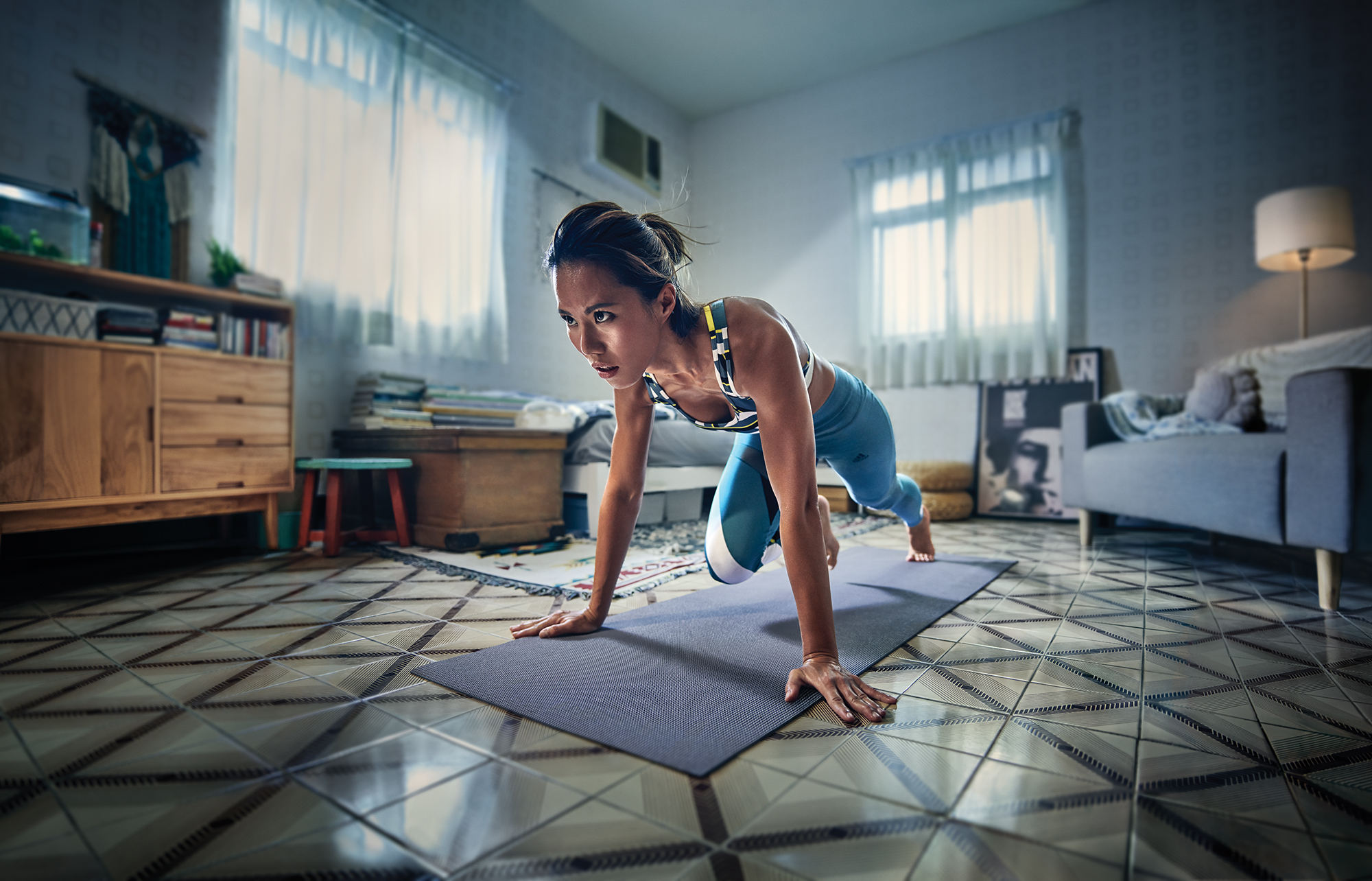 Athletic Asian woman in sports bra and leggings does mountain climber exercise on a yoga mat in a living room with tile floor.