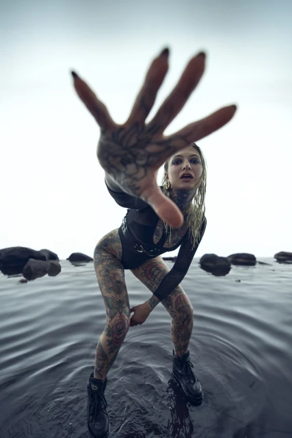 Tattooed woman in a black bodysuit reaches towards the camera while standing in shallow water with rocks, under a bright sky.