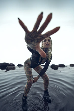 Tattooed woman in a black bodysuit reaches towards the camera while standing in shallow water with rocks, under a bright sky.