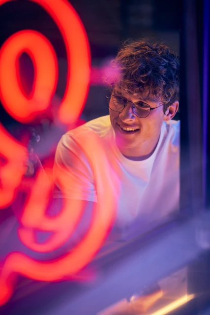 Portrait of a smiling young adult man with curly hair and glasses, standing indoors in front of a blurry bright red neon light.