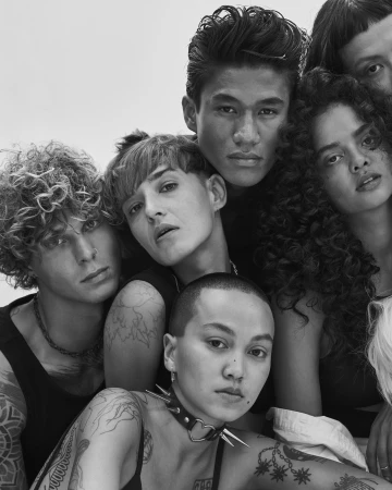 Black and white portrait of a diverse group of seven people, some tattooed, with varying hairstyles, closely framed in an indoor setting.
