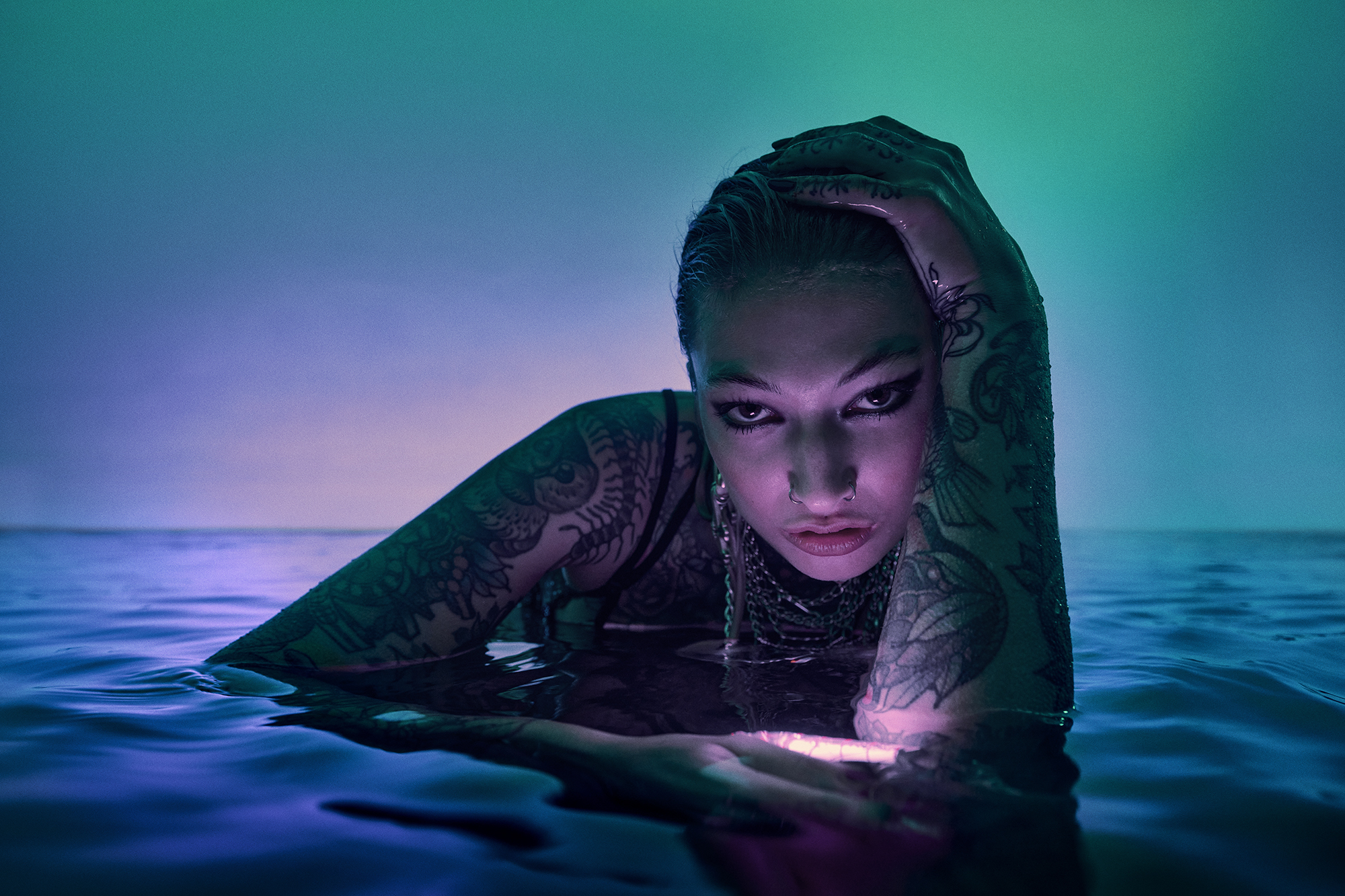Woman in body of water with teal and blue lighting and tattooed arms. Close up portrait view of person in water, artistic effect.