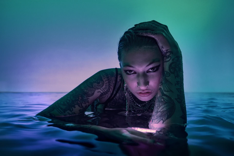 Woman in body of water with teal and blue lighting and tattooed arms. Close up portrait view of person in water, artistic effect.